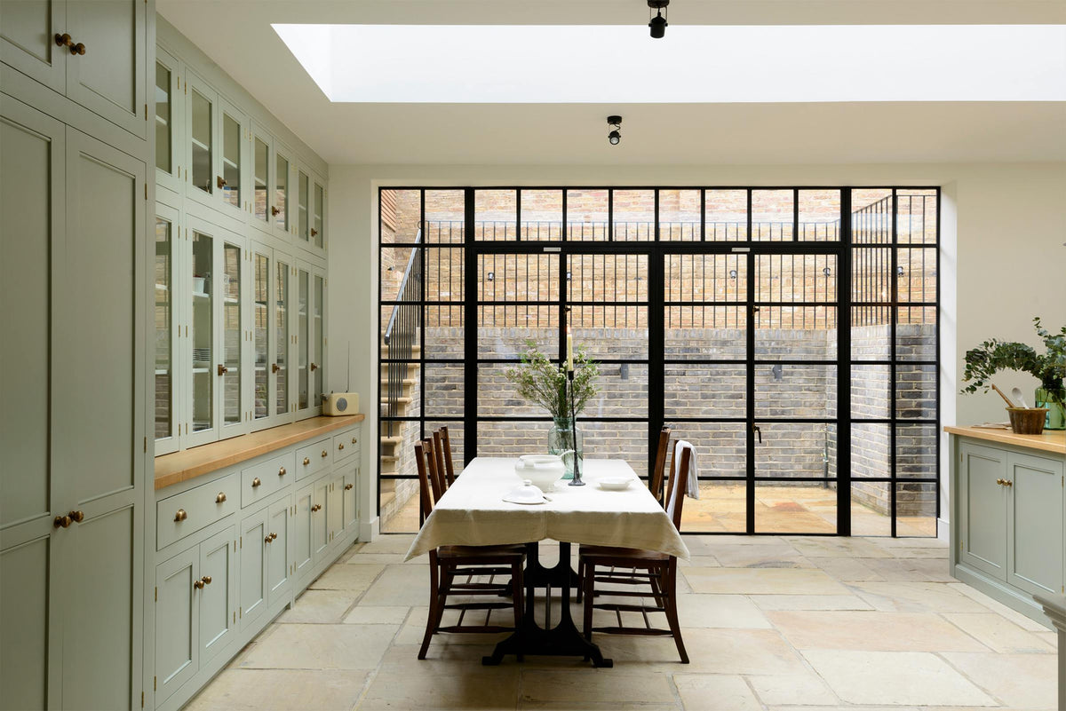 This bright and spacious classic English kitchen features a full-height cupboard, a long pantry run with glazed counter cupboards, and a solid oak worktop. Adorned with a vintage cream radio, an aqua-green vase filled with a floral arrangement, and vintage wooden furniture, it creates a delightful mix of simplicity and charismatic personality.