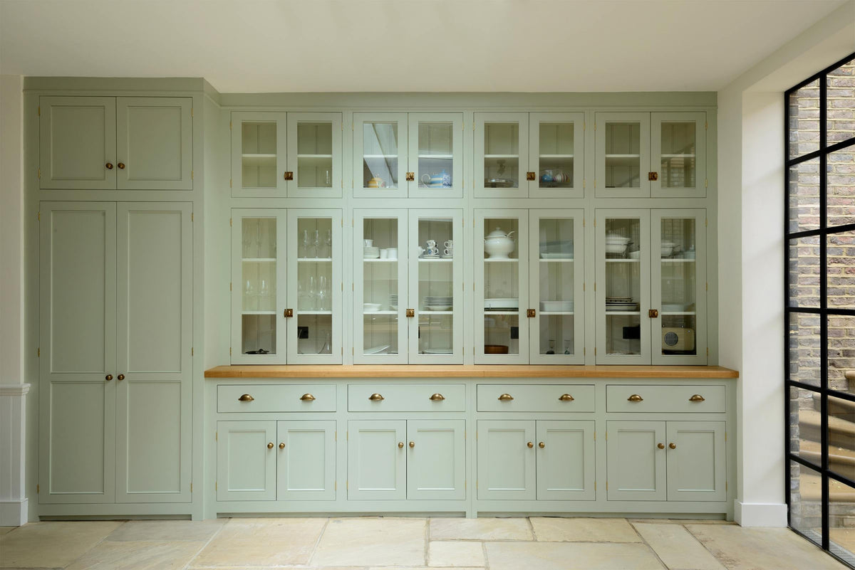 This bright and spacious classic English kitchen features a full-height cupboard, a long pantry run with glazed counter cupboards, and a solid oak worktop. Adorned with a vintage cream radio, an aqua-green vase filled with a floral arrangement, and vintage wooden furniture, it creates a delightful mix of simplicity and charismatic personality.