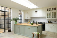 This bright and spacious classic English kitchen features a long white cooker run with glazed wall cupboards and a pale-blue kitchen island that includes a single Belfast sink, an aged brass Ionian tap, and a solid oak worktop. Adorned with metal embellishments like industrial pendant lights, a silver refrigerator, a jade-green vase filled with a floral arrangement, and vintage wooden furniture, it creates a wonderful mix of simplicity and charismatic personality.