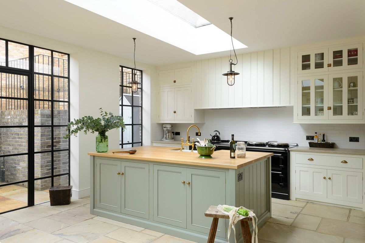 This bright and spacious classic English kitchen features a long white cooker run with glazed wall cupboards and a pale-blue kitchen island that includes a single Belfast sink, an aged brass Ionian tap, and a solid oak worktop. Adorned with metal embellishments like industrial pendant lights, a silver refrigerator, a jade-green vase filled with a floral arrangement, and vintage wooden furniture, it creates a wonderful mix of simplicity and charismatic personality.