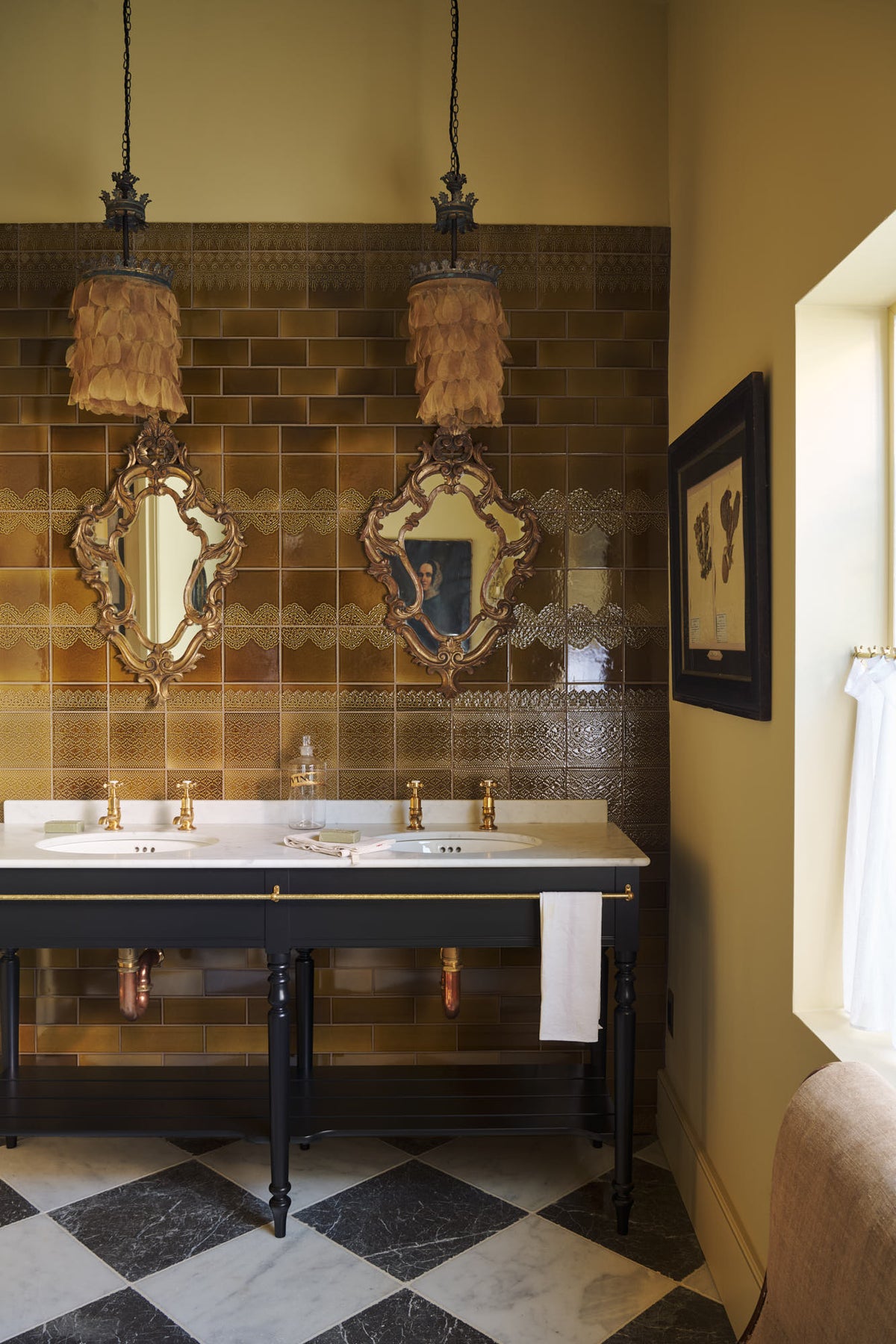 A beautiful butter-yellow powder room, you’ll find a black twin Victorian washstand, tall sepia lace market tiles, and a Parisian chequered marble floor. Paired with antique mirrors, pendant lights, vintage paintings, and framed pressed botanical artworks, the room exudes a glamorous, vintage charm.