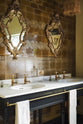 A close-up and oblique view of a butter-yellow powder room, you’ll find a black twin Victorian washstand, and tall sepia lace market tiles. Paired with antique mirrors, pendant lights, towels, and framed pressed botanical artworks, the room exudes a glamorous, vintage charm.