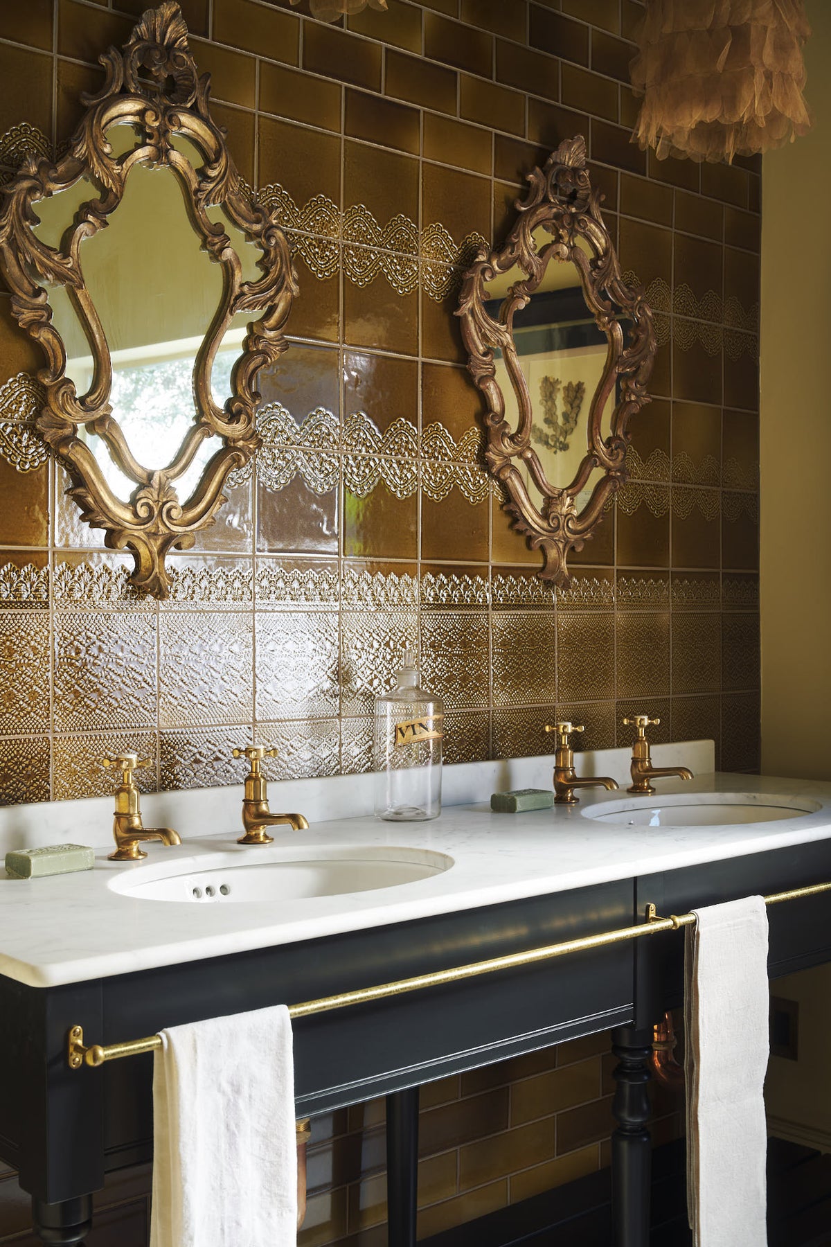 A close-up and oblique view of a butter-yellow powder room, you’ll find a black twin Victorian washstand, and tall sepia lace market tiles. Paired with antique mirrors, pendant lights, towels, and framed pressed botanical artworks, the room exudes a glamorous, vintage charm.