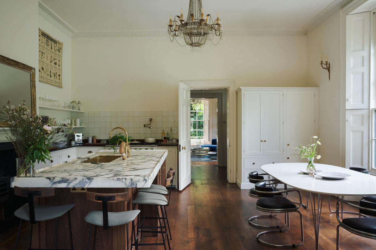 A classic English kitchen in white linen features white and cream splashback tiles, a cooker with a polished sapele worktop, and a kitchen island topped with Arabescato marble. It includes a polished brass sink, an aged brass Ionian tap, a scientific illustration of vegetable species, two glasses of local wildflowers, and a metal-and-crystal chandelier.