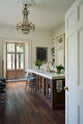Looking through the door of a grand yet simple linen-white classic English kitchen, one sees an Arabescato marble kitchen island, a polished brass sink, and an aged brass Ionian tap. The space is simply decorated with books, glass vases filled with wildflowers from the area, and a large metal-and-crystal chandelier.