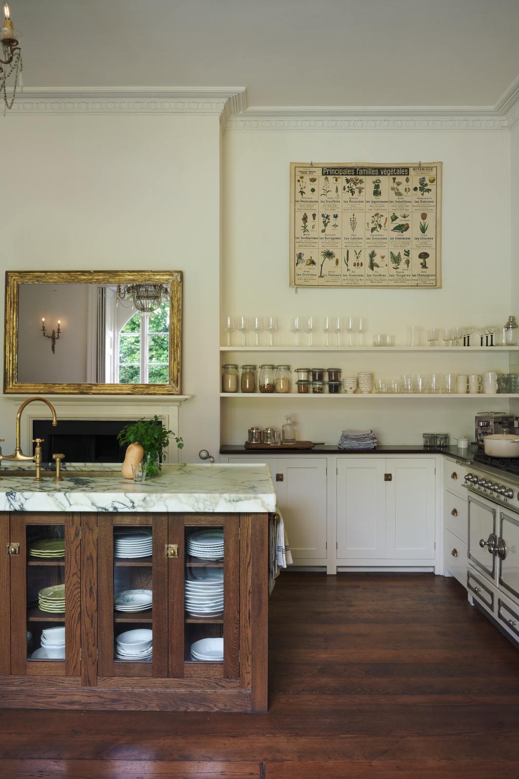 A grand yet simple white linen classic English kitchen, featuring white and cream splashback tiles, a cooker run with a polished sapele worktop, and a kitchen island topped with Arabescato marble, a polished brass sink, and an aged brass Ionian tap. The space is decorated with a large scientific-style illustration of different vegetable species and a large metal-and-crystal chandelier.