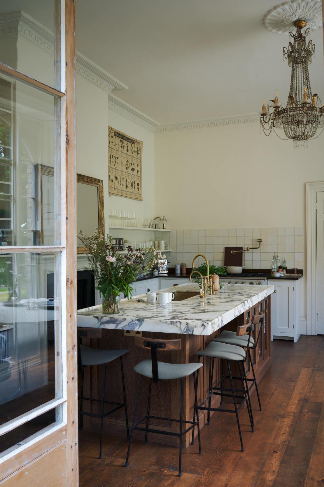 Looking through the door of a grand yet simple linen-white classic English kitchen, one sees white and cream splashback tiles, a cooker run with a polished sapele worktop, and a kitchen island topped with Arabescato marble, a polished brass, and an aged brass Ionian tap. The space is simply decorated with books, glass vases filled with wildflowers from the area, and a large metal-and-crystal chandelier.