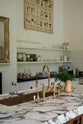 A closer look at the kitchen island reveals a surface topped with Arabescato marble, a polished brass sink, and an aged brass Ionian tap—all part of a grand yet simple linen-white classic English kitchen. The space is simply decorated with a large scientific-style illustration of various vegetable species, a garnish of parsley beside a butternut squash, and a gold-framed mirror above an unused fireplace.