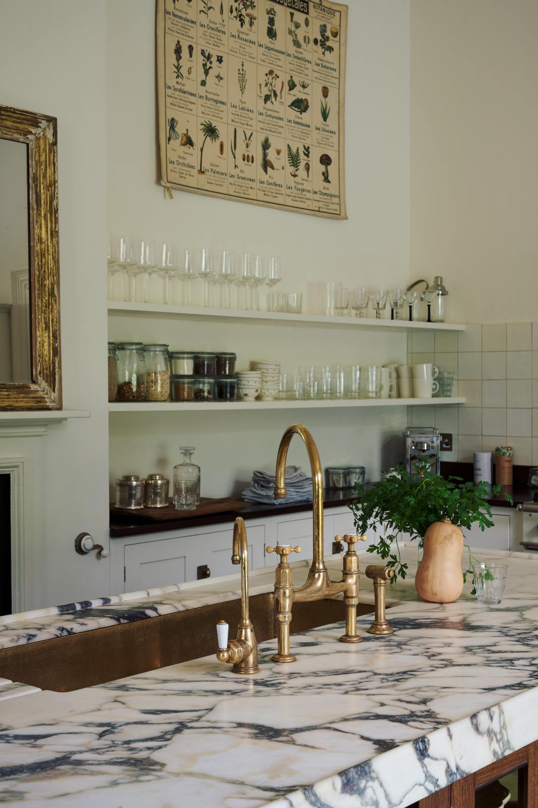 A closer look at the kitchen island reveals a surface topped with Arabescato marble, a polished brass sink, and an aged brass Ionian tap—all part of a grand yet simple linen-white classic English kitchen. The space is simply decorated with a large scientific-style illustration of various vegetable species, a garnish of parsley beside a butternut squash, and a gold-framed mirror above an unused fireplace.