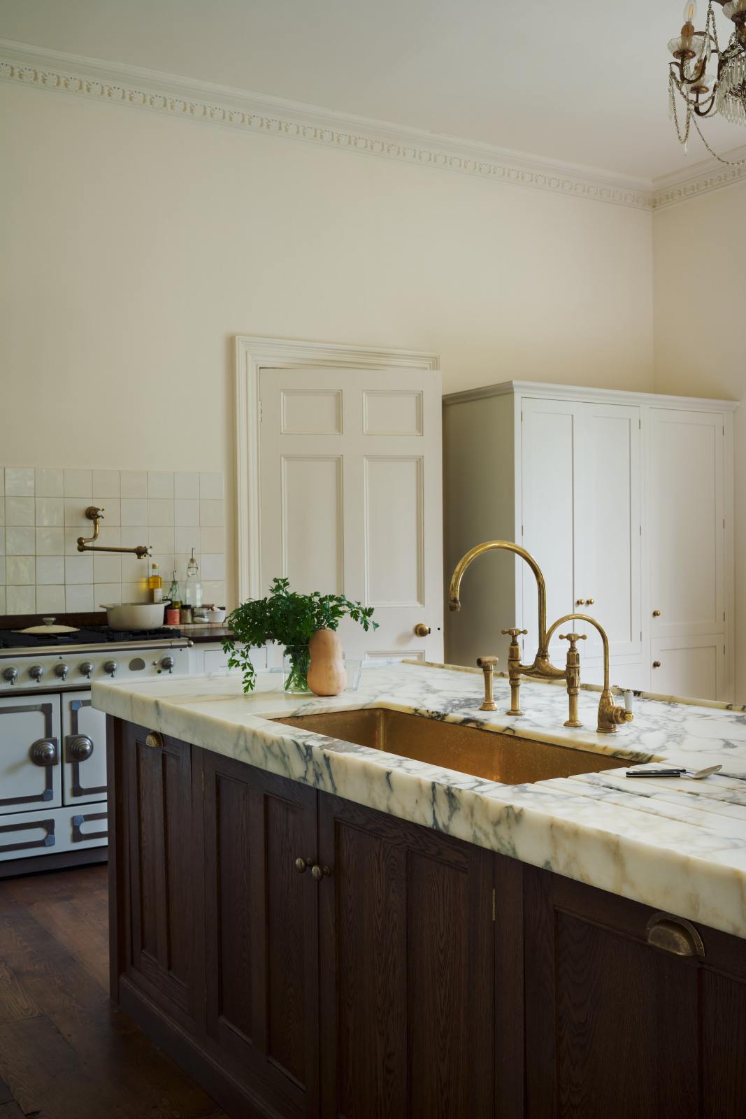 A closer look at the kitchen island reveals a surface topped with Arabescato marble, a polished brass sink, and an aged brass Ionian tap—all part of a grand yet simple linen-white classic English kitchen. The space is decorated with a garnish of parsley beside a butternut squash, and a large metal-and-crystal chandelier.