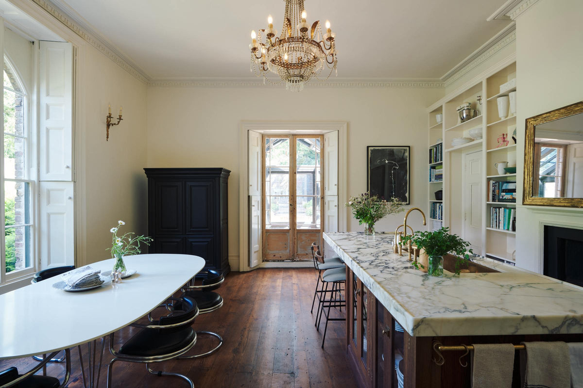In a grand yet simple linen-white classic English kitchen, one sees an Arabescato marble kitchen island, a polished brass sink, an aged brass Ionian tap, a large dining table with metal-framed chairs upholstered in black cushions, and a vintage French door leading to the garden. The space is simply decorated with books, glass vases filled with wildflowers from the area, and a large metal-and-crystal chandelier.