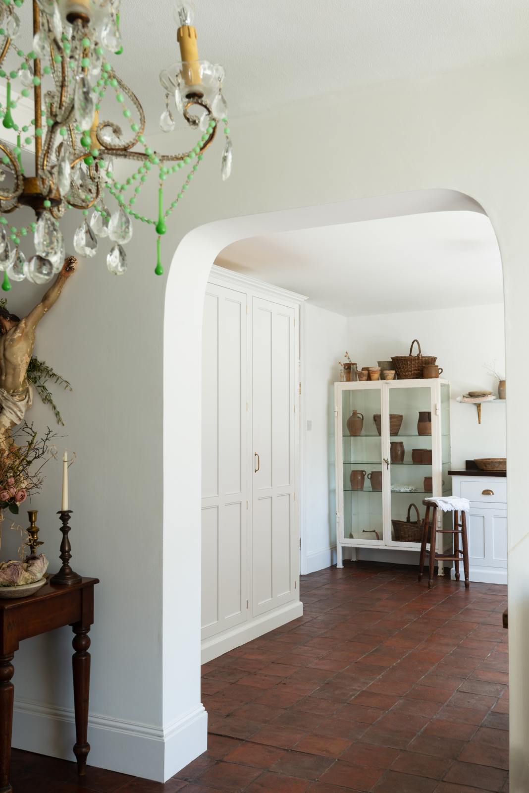 An oblique view of an arched entrance to a simple and functional linen white scullery room with a modest sink run with polished sapele, and full-height cabinets. The warm reds and browns of the terracotta square tiles, vintage furniture, ceramics, and a deVOL laundry maid serve the room with a purpose and beautiful aesthetics.