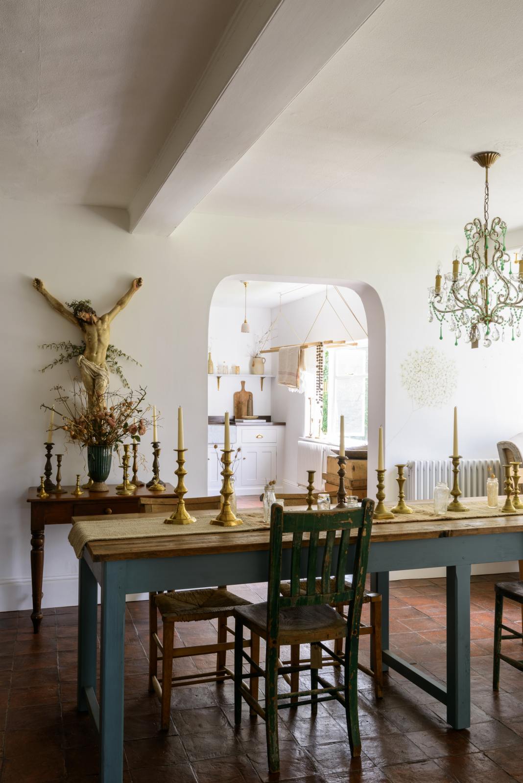 A gorgeous dining room features a teal Millhouse refectory table with vintage wooden chairs, furniture, a large variety of brass candlesticks, a crystal chandelier and a Jesus statue on the wall. And through the open archway is a simple and functional linen white scullery room with a modest sink run with polished sapele, a large single Carrara marble sink, and a deVOL laundry maid.