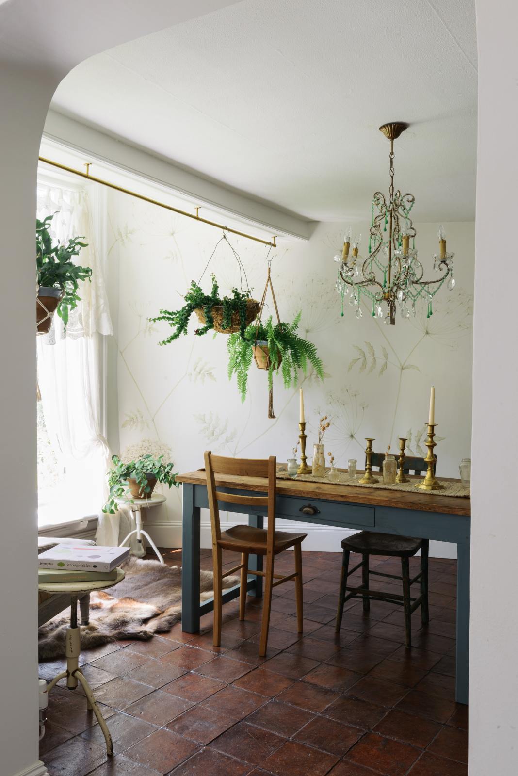 Looking through an open archway is a beautiful dining room with a teal Millhouse refectory table, vintage wooden chairs, an array of brass candlesticks, a crystal chandelier, hanging plants and an intricate wild flower illustration wallpaper.