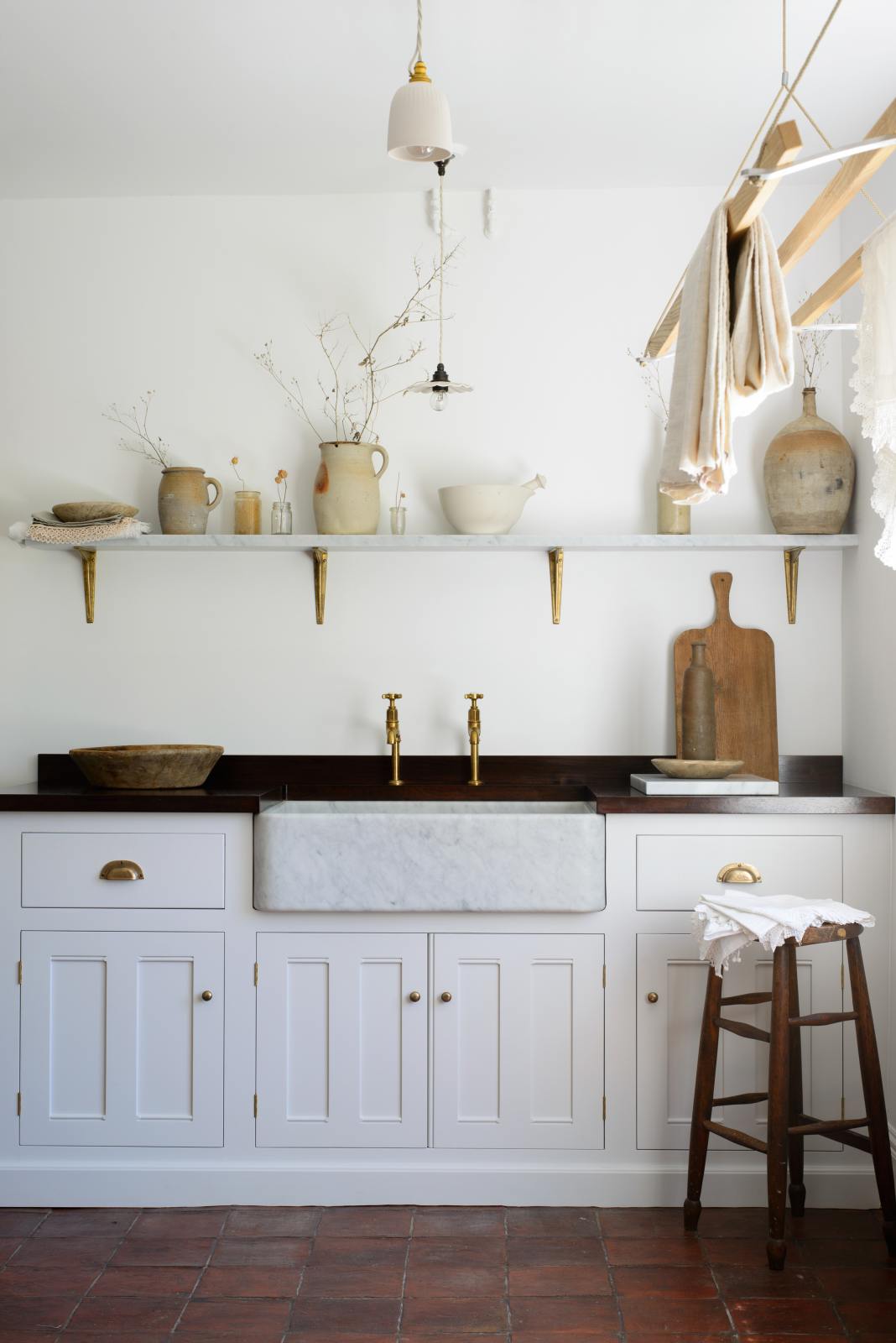 The simple and functional linen white scullery room with a modest sink run with polished sapele, a large single Carrara marble sink, and aged brass Mayan taps. The warm reds and browns of the terracotta square tiles, vintage furniture, ceramics, woven baskets and a deVOL laundry maid serve the room with both a purpose and beautiful aesthetics.