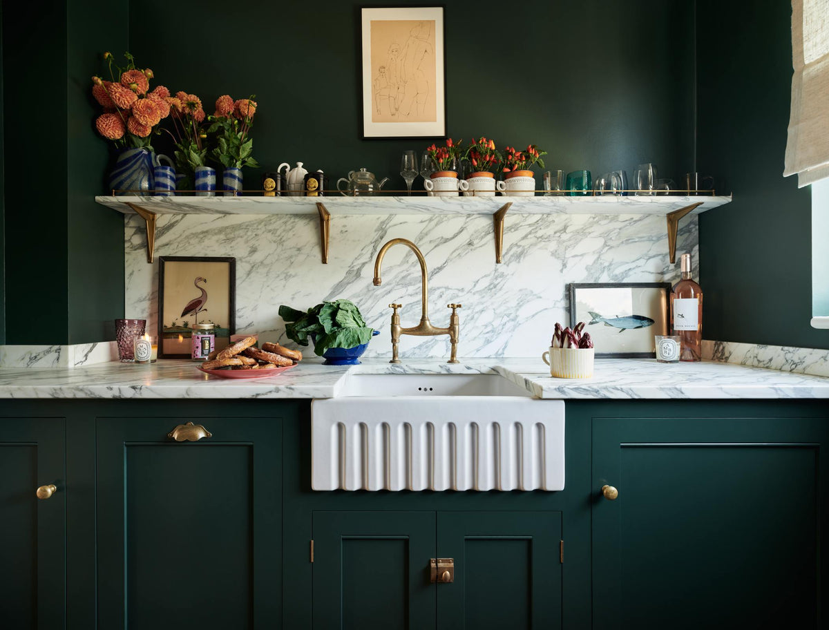 This charming Bakehouse green English kitchen features an Arabescato worktop, splashback, and counter shelf, along with a single fluted Belfast sink and an aged brass Ionian tap. Decorated with colourful bric-a-brac, framed illustrations, pretzels, washed lettuce in a blue strainer, and red chicory in a large mug, it perfectly blends functionality with personality.