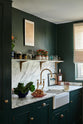 An angled view of a small but luxurious Bakehouse green classic English kitchen featuring a sink run with an Arabescato worktop, splashback and counter shelf, a single fluted Belfast sink and an aged brass Ionian tap. Beautifully decorated with all sorts of colourful and intricate bric-a-brac, framed illustrations, a plate of pretzels, washed lettuce in a blue strainer, and red chicory in a large mug, giving the kitchen functionality and personality.