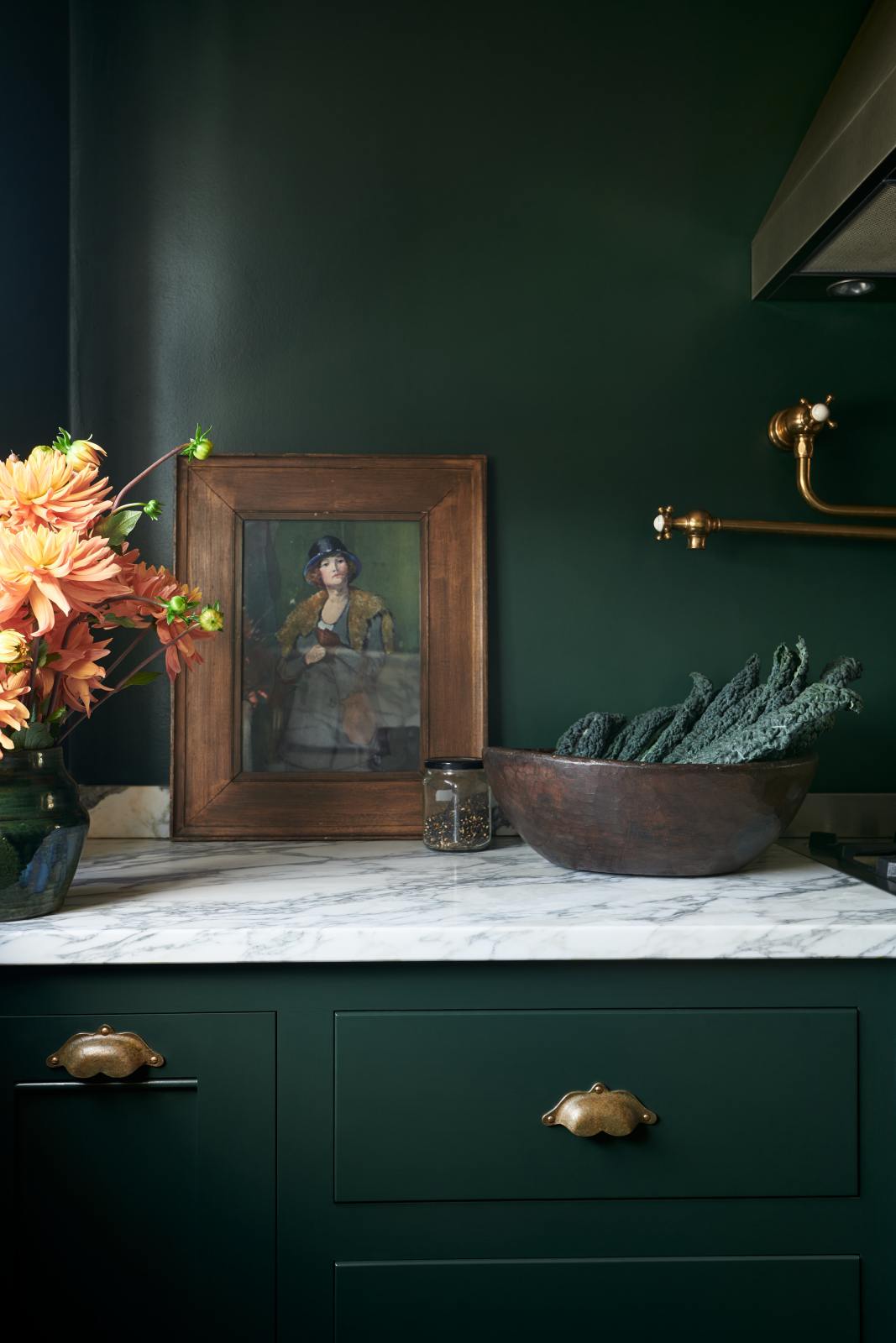 A quiet corner of a small but luxurious Bakehouse green classic English kitchen features a cooker run with an Arabescato worktop and a brass pot filler tap. Beautifully decorated with peach and yellow flowers in an emerald vase, a framed portrait painting of a woman, a wooden bowl of washed lettuce, and a jar of spices, giving the kitchen functionality and personality.