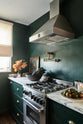 An angled view of a small, luxurious Bakehouse green classic English kitchen features Arabescato worktops, a brass pot filler tap, and a silver gas cooker with a canopy. It’s adorned with peach and yellow flowers in an emerald vase, a portrait of a woman, a bowl of washed lettuce and green tomatoes, and a jar of loose tea, adding both functionality and personality.