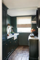A small but luxurious Bakehouse green classic English kitchen features a sink and cooker run with an Arabescato worktop, splashback and counter shelf, a single fluted Belfast sink and a full-height cabinet. It’s adorned with all sorts of colourful and intricate bric-a-brac, framed illustrations, a plate of pretzels, washed lettuce, and peachy yellow flowers, giving the kitchen functionality and personality.