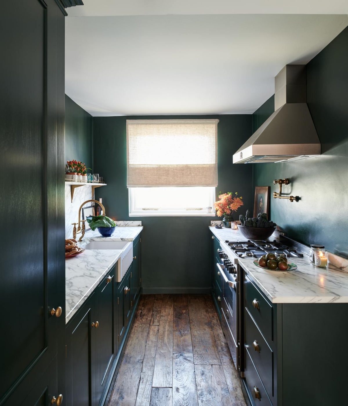 A small but luxurious Bakehouse green classic English kitchen features a sink and cooker run with an Arabescato worktop, splashback and counter shelf, a single fluted Belfast sink and a full-height cabinet. It’s adorned with all sorts of colourful and intricate bric-a-brac, framed illustrations, a plate of pretzels, washed lettuce, and peachy yellow flowers, giving the kitchen functionality and personality.