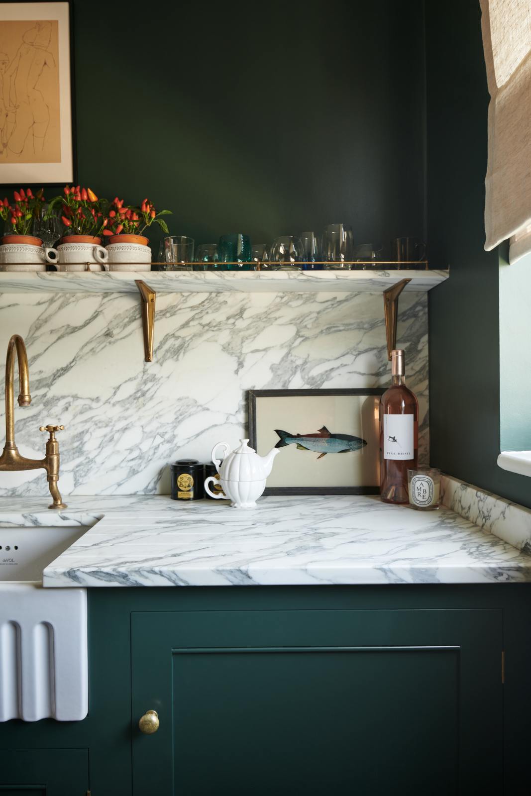 A quiet corner of a small but luxurious Bakehouse green classic English kitchen features a sink run with an Arabescato worktop, splashback and counter shelf, a single fluted Belfast sink and an aged brass Ionian tap.  It’s adorned with decorated with all sorts of colourful and intricate bric-a-brac, framed illustrations, a variety of drinking glasses, wine and small pots of red peppers, giving the kitchen both functionality and personality.