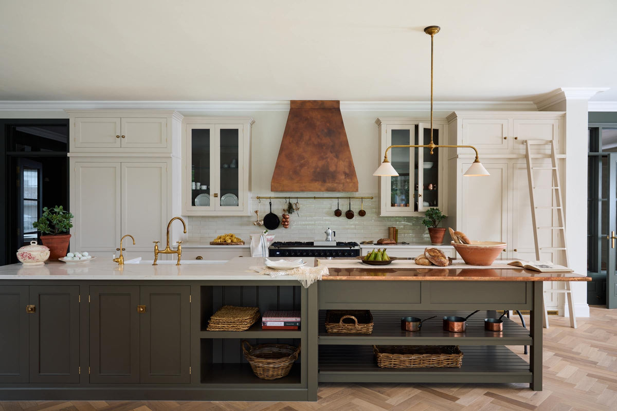 A grand, earth-toned classic kitchen features a Silestone worktop cooker run, glazed wall cupboards, a full-height pantry, a copper canopy, and a large island with a sink, topped with Silestone on one side and copper on the other. Adorned with warm colours such as an aged brass hanging rail, deVOL Heirloom gaselier lights, textured woven baskets, vintage ceramics and loaves of bread, it makes a spectacular use of space but is also down-to-earth.