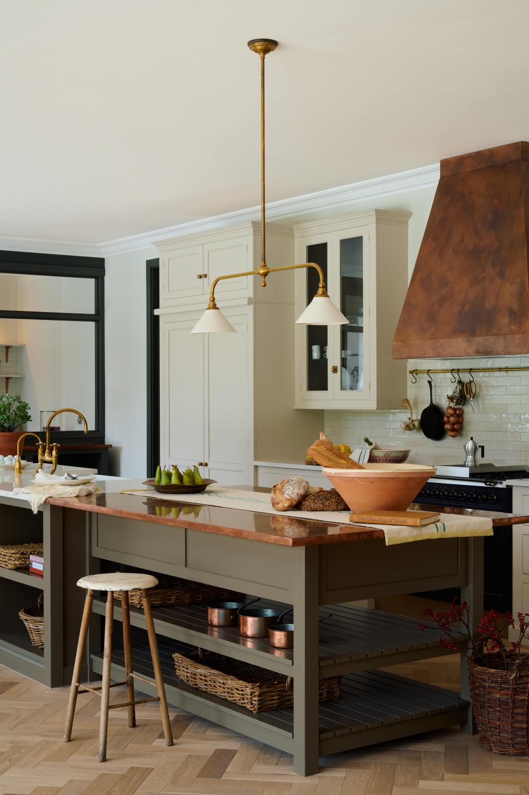 An oblique view of a grand, earth-toned classic kitchen features a Silestone worktop cooker run, glazed wall cupboards, a full-height pantry, a copper canopy, and a large island with a sink, topped with Silestone on one side and copper on the other. Adorned with warm colours such as an aged brass hanging rail, deVOL Heirloom gaselier lights, textured woven baskets, vintage ceramics and loaves of bread, it makes a spectacular use of space but also down-to-earth.