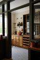 An angled view of a large Haberdasher’s walk-in pantry, featuring Tambour base cabinets, open drawers, glazed countertop cupboards, a Silestone worktop, and shelves. Adorned with natural materials such as white porcelain splashback tiles, classic brass shelf brackets, an intricately patterned tiled floor, wooden chopping boards, vintage ceramics, vegetables, and loaves of bread; it makes spectacular use of the space while remaining down-to-earth.