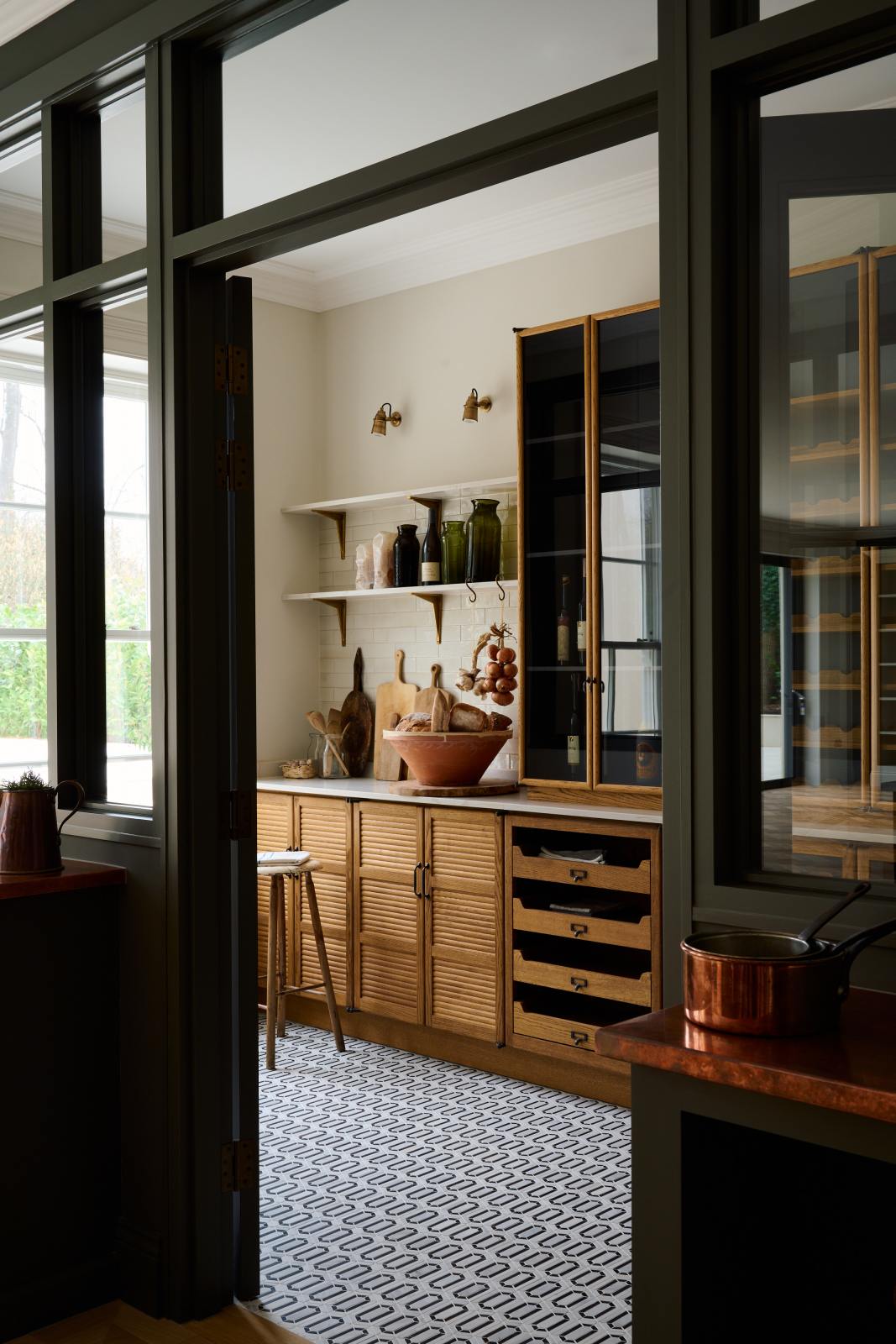 An angled view of a large Haberdasher’s walk-in pantry, featuring Tambour base cabinets, open drawers, glazed countertop cupboards, a Silestone worktop, and shelves. Adorned with natural materials such as white porcelain splashback tiles, classic brass shelf brackets, an intricately patterned tiled floor, wooden chopping boards, vintage ceramics, vegetables, and loaves of bread; it makes spectacular use of the space while remaining down-to-earth.