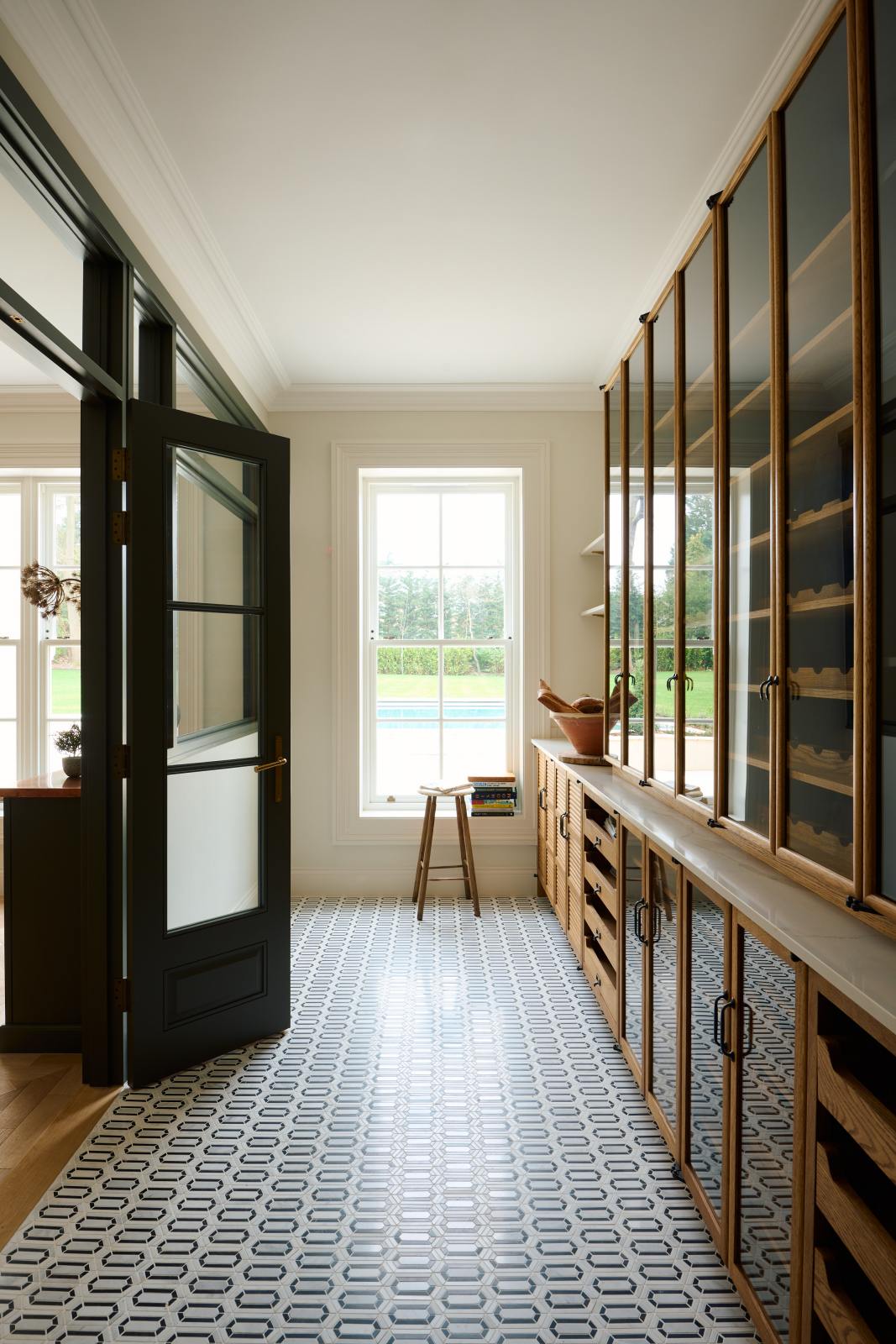 A large Haberdasher’s walk-in pantry, featuring Tambour base cabinets, open drawers, glazed countertop cupboards, a Silestone worktop, and shelves. Adorned with natural materials such as a white vintage stool, books, an intricately patterned tiled floor, wooden chopping boards, vintage ceramics, and loaves of bread, it makes spectacular use of the space while remaining down-to-earth.