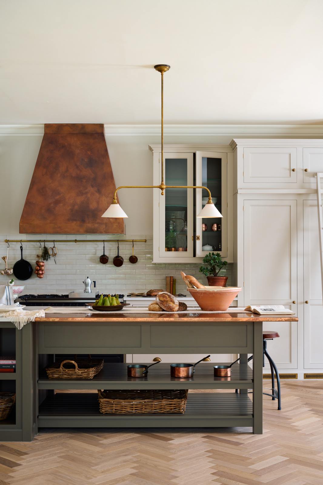 A grand, earth-toned classic kitchen features a Silestone worktop cooker run, glazed wall cupboards, a full-height pantry, a copper canopy, and a large island with a sink, topped with Silestone on one side and copper on the other. Adorned with warm colours such as an aged brass hanging rail, deVOL Heirloom gaselier lights, textured woven baskets, vintage ceramics and loaves of bread, it makes a spectacular use of space but is also down-to-earth.