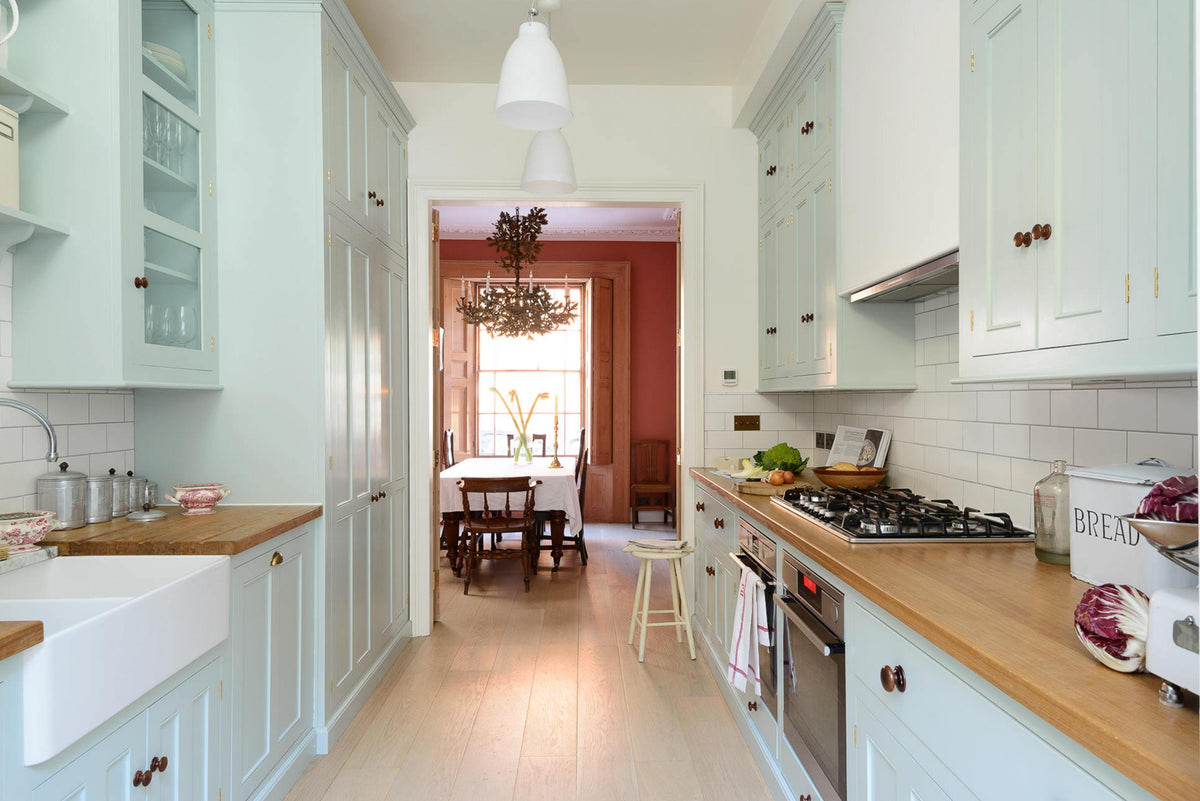 This pale-blue classic English galley kitchen features a cooker and sink run with a solid oak worktop, a double Belfast sink, a chrome Ionian tap, wall cupboards, a canopy, and shelves. Adorned with a vintage scale, a bread tin, artisan ceramics, silver containers, and colourful vegetables, it creates a humble and practical kitchen that embraces vintage 80s aesthetics.