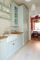An angled view of a pale-blue classic English galley kitchen features a sink run with a solid oak worktop, a double Belfast sink, a chrome Ionian tap, glazed wall cupboards, pantry cupboards, and shelves. Adorned with artisan ceramics, vintage containers, and simple platters, it creates a humble and practical kitchen that embraces vintage 80s aesthetics.