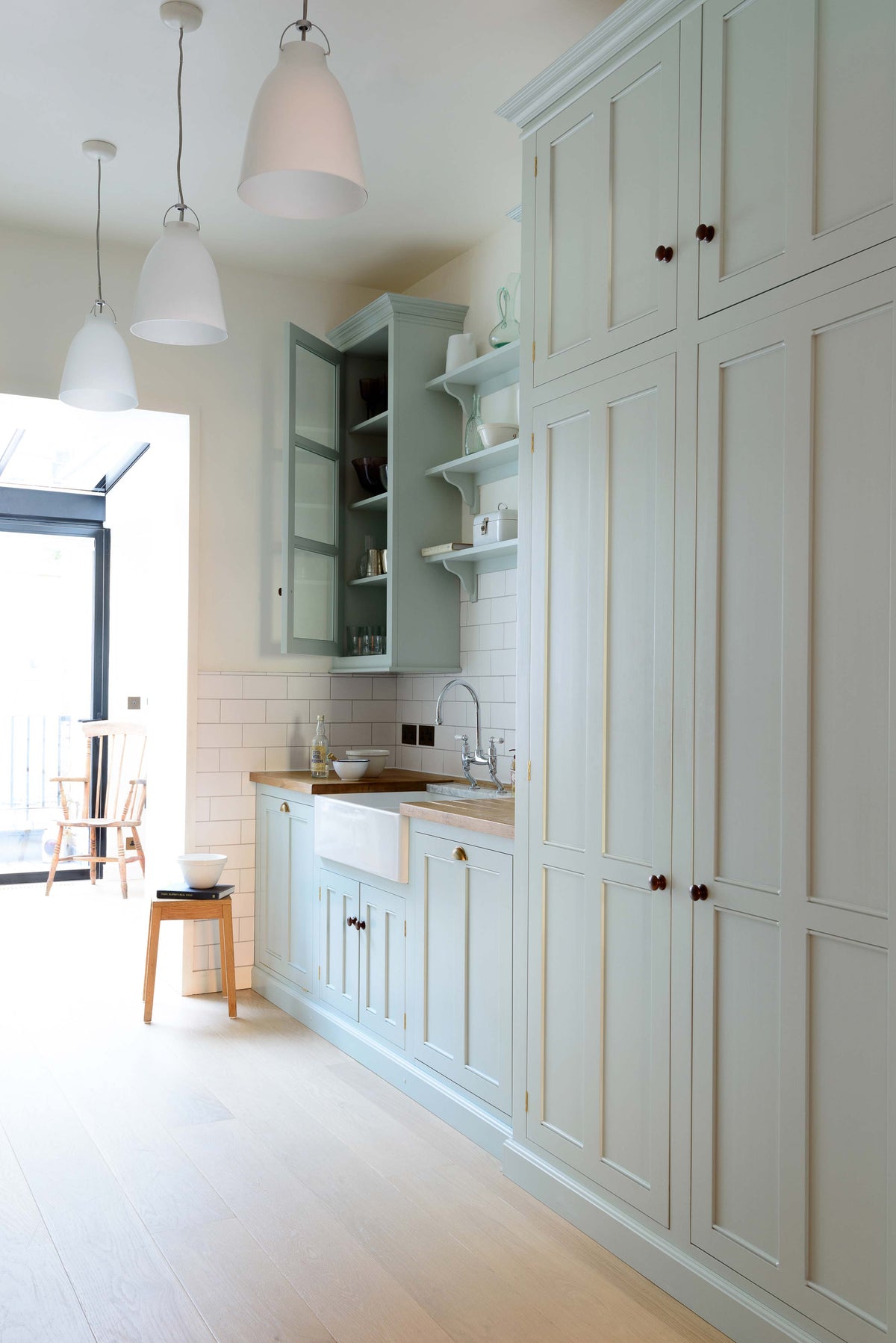 An angled view of a pale-blue classic English galley kitchen features a sink run with a solid oak worktop, a double Belfast sink, a chrome Ionian tap, glazed wall cupboards, pantry cupboards, and shelves. Adorned with artisan ceramics, vintage containers, and simple platters, it creates a humble and practical kitchen that embraces vintage 80s aesthetics.