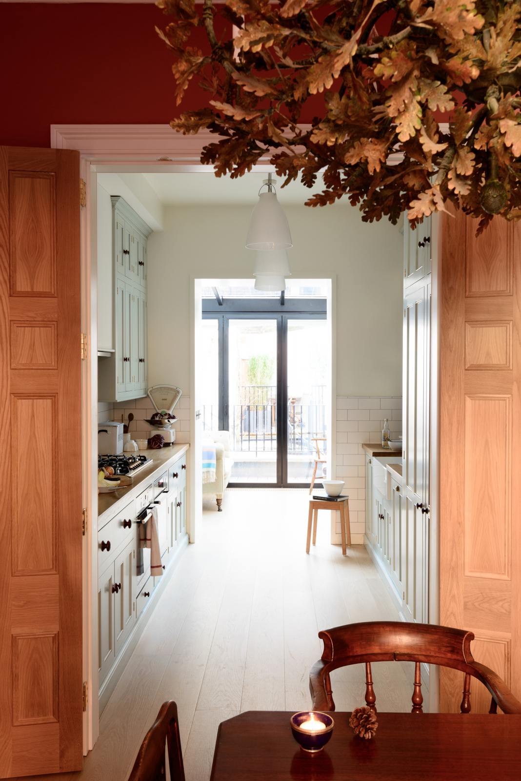 Beyond a cosy wooden dining room featuring a solid oak dining table, chairs, and a detailed oak chandelier, you will find a pale-blue classic English galley kitchen. This leads into a bright, lounging area, adorned with floor-to-ceiling glass windows, allowing plenty of natural light.