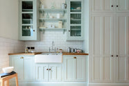 This pale-blue classic English galley kitchen features a sink run with a solid oak worktop, a double Belfast sink, a chrome Ionian tap, glazed wall cupboards, pantry cupboards, and shelves. Adorned with artisan ceramics, vintage containers, and simple platters, it creates a humble and practical kitchen that embraces vintage 80s aesthetics.