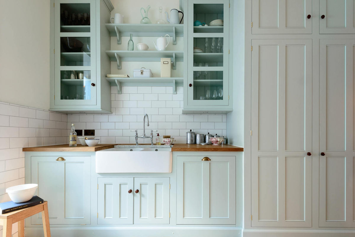 This pale-blue classic English galley kitchen features a sink run with a solid oak worktop, a double Belfast sink, a chrome Ionian tap, glazed wall cupboards, pantry cupboards, and shelves. Adorned with artisan ceramics, vintage containers, and simple platters, it creates a humble and practical kitchen that embraces vintage 80s aesthetics.