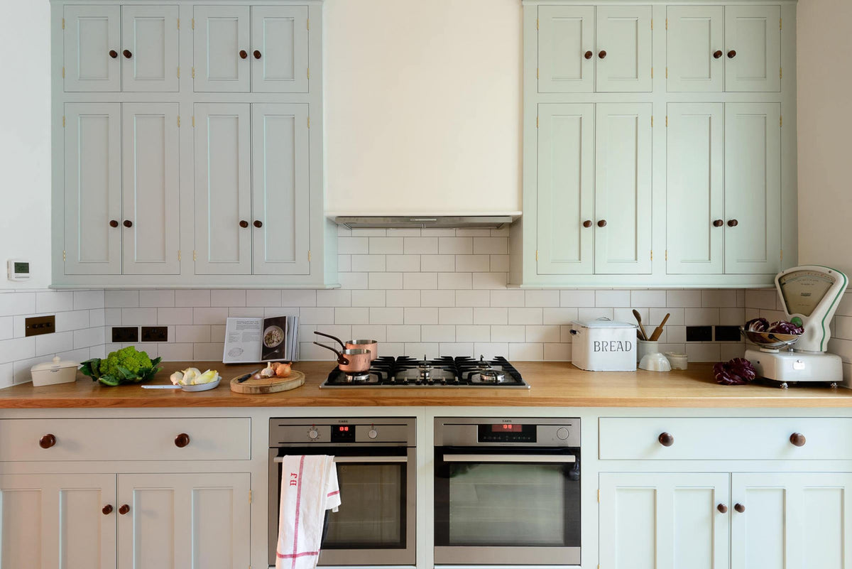 This pale-blue classic English galley kitchen features a cooker run with a solid oak worktop, a canopy, a silver gas cooker, and two ovens. Adorned with a vintage scale, a bread tin, simple ceramics, copper pans, and colourful vegetables, it creates a humble and practical kitchen that embraces vintage 80s aesthetics.