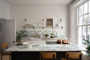 A humble yet elegant off-white Classic English kitchen features a sink run with honed Arabescato marble worktop and splashback, a pantry blue kitchen island with a cooker, mini creamware wall lights, and plumber’s stools. Adorned with artisan ceramics, brass pans, a vintage painting, fresh greens, and grains, creating an intimate social and practical kitchen space.