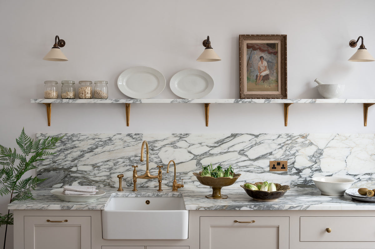 A close-up of a humble yet elegant off-white Classic English kitchen features a sink run with honed Arabescato marble worktop and splashback, a marble shelf, mini creamware wall lights, and an aged brass Ionian tap. Adorned with artisan ceramics, a vintage painting, fresh greens, and grains, creating an intimate social and practical kitchen space.