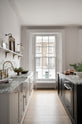 A humble yet elegant off-white Classic English kitchen features a sink run with honed Arabescato marble worktop and splashback, a pantry blue kitchen island with a cooker, and mini creamware wall lights. Adorned with artisan ceramics, brass pans, a vintage painting, fresh greens, and grains, creating an intimate social and practical kitchen space.