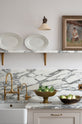 A close-up of a humble yet elegant off-white Classic English kitchen features a sink run with honed Arabescato marble worktop and splashback, a marble shelf, mini creamware wall lights, and an aged brass Ionian tap. Adorned with artisan ceramics, a vintage painting, and fresh greens, creating an intimate social and practical kitchen space.