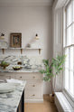 A quiet corner of a humble yet elegant off-white Classic English kitchen features a sink run with honed Arabescato marble worktop and splashback, a pantry blue kitchen island with a cooker, and mini creamware wall lights. Adorned with artisan ceramics, a vintage painting, fresh greens, and grains, creating an intimate social and practical kitchen space.