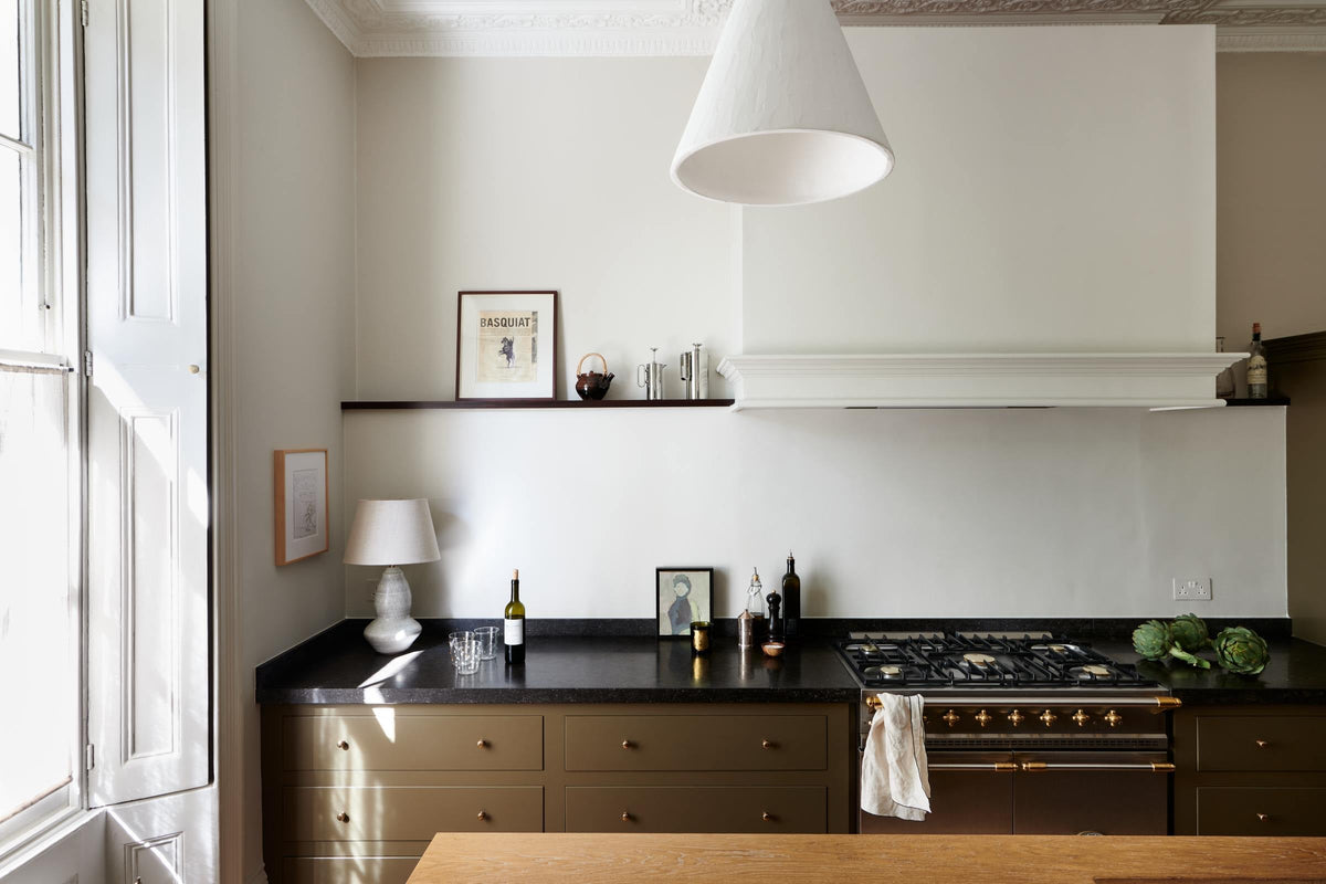 An elegant olive-brown classic kitchen features a cooker run topped with Belgian blue limestone, a kitchen island with a sink and a solid oak worktop, a shaker canopy, and a built-in shelf. The tall-ceilinged room is decorated with slender bottles of wine and vinegar, a pepper grinder, a vibrant green artichoke, and small, delicate framed illustrations.