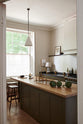 Looking through a white open door, one sees an elegant olive-brown classic kitchen featuring a cooker run topped with Belgian blue limestone, a Shaker canopy, and a built-in shelf. At the centre is a kitchen island with a sink, a solid oak worktop, an aged brass Ionian tap, and a breakfast nook with wooden stools at the far edge. The tall-ceilinged room is decorated with slender bottles of wine and vinegar, a pepper grinder, a vibrant green artichoke, and small, delicate framed illustrations.