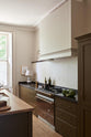 An oblique view of an elegant olive-brown classic kitchen features a cooker run topped with Belgian blue limestone, a kitchen island with a sink and a solid oak worktop, a shaker canopy, and a built-in shelf. The tall-ceilinged room is decorated with slender bottles of wine and vinegar, a pepper grinder, a vibrant green artichoke, and small, delicate framed illustrations.