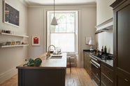 Looking out through the wide windows of an elegant olive-brown classic kitchen, one sees a space framed by a cooker run topped with Belgian blue limestone and a kitchen island with a sink, a solid oak worktop, an aged brass Ionian tap, and wooden stools at the far edge. The tall-ceilinged room is decorated with slender bottles of wine and vinegar, a pepper grinder, a vibrant green artichoke, and small, delicate framed illustrations.