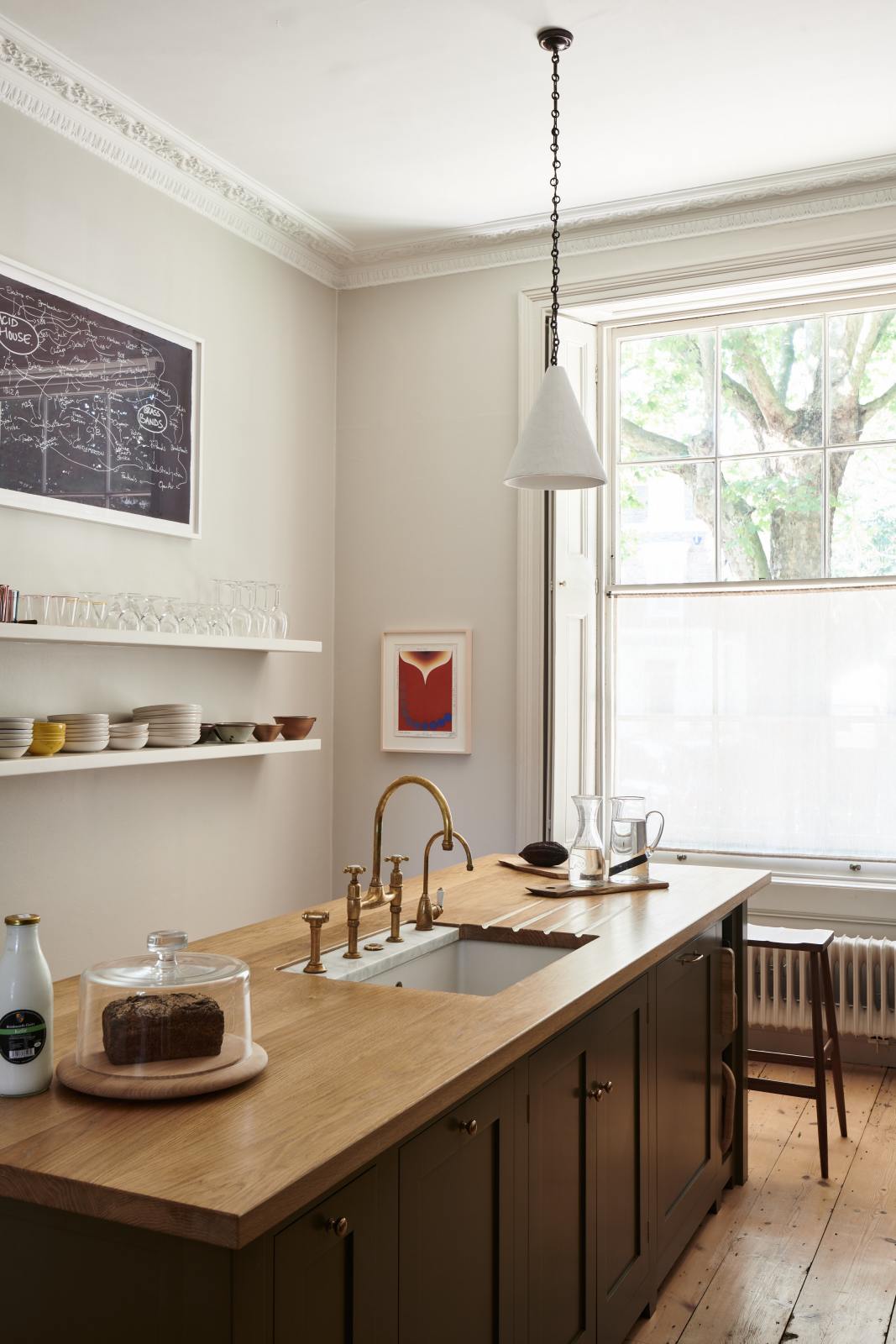 An oblique view of an elegant olive-brown classic kitchen features a kitchen island with a sink, a solid oak worktop, an aged brass Ionian tap, and a breakfast nook with wooden stools at the far edge. Long shelves line the wall, stacked with ceramic bowls, wine glasses, cocktail glasses, and drinking glasses.