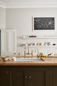 An elegant olive-brown classic kitchen features a kitchen island with a sink, a solid oak worktop, an aged brass Ionian tap, and a breakfast nook with wooden stools at the far edge. Long shelves line the wall, stacked with ceramic bowls, wine glasses, cocktail glasses, and drinking glasses. The tall-ceilinged room is decorated with slender water pitchers, glass milk, a bag of oranges, and big and colourful framed illustrations.